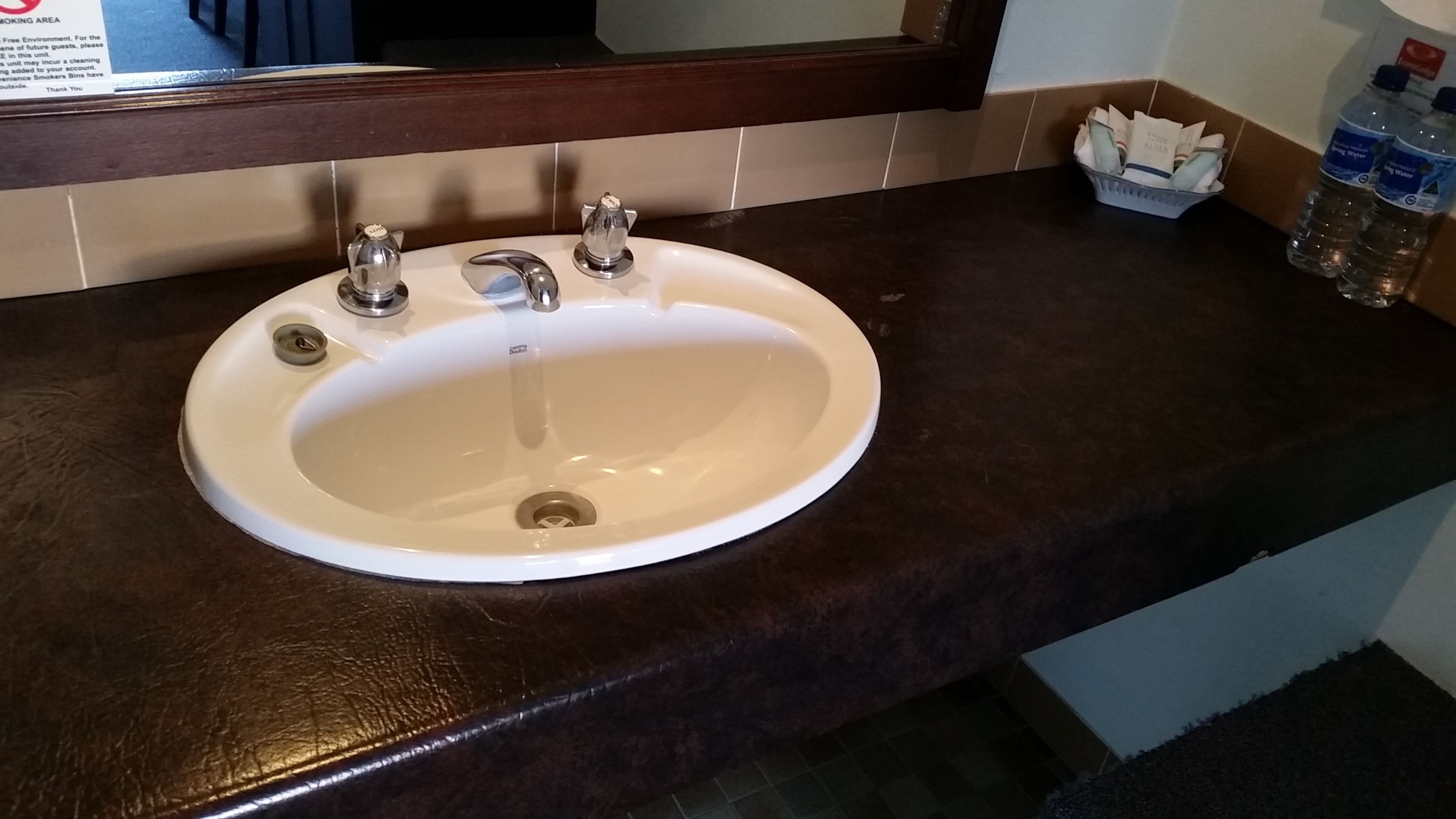 A hotel bathroom sink with a mirror above it, a towel dispenser, and a bottle of water on the counter.
