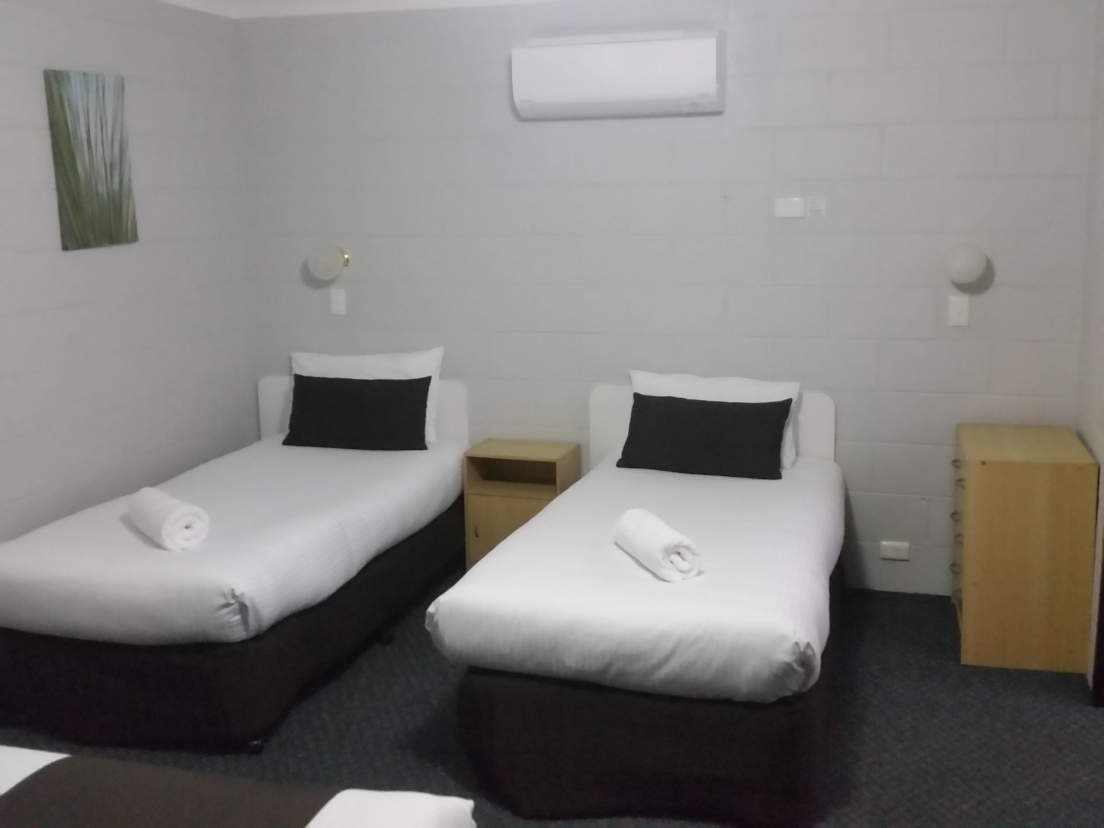 A room with two single beds, each with a pillow and a rolled towel. There is a small wooden nightstand and a wooden cabinet.
