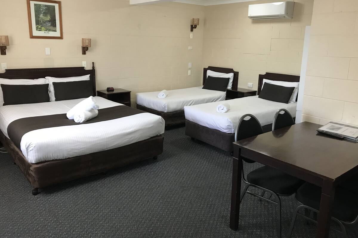 A hotel room with three beds, a desk, and a chair. There is a framed picture on the wall and an air conditioning unit mounted on the wall.