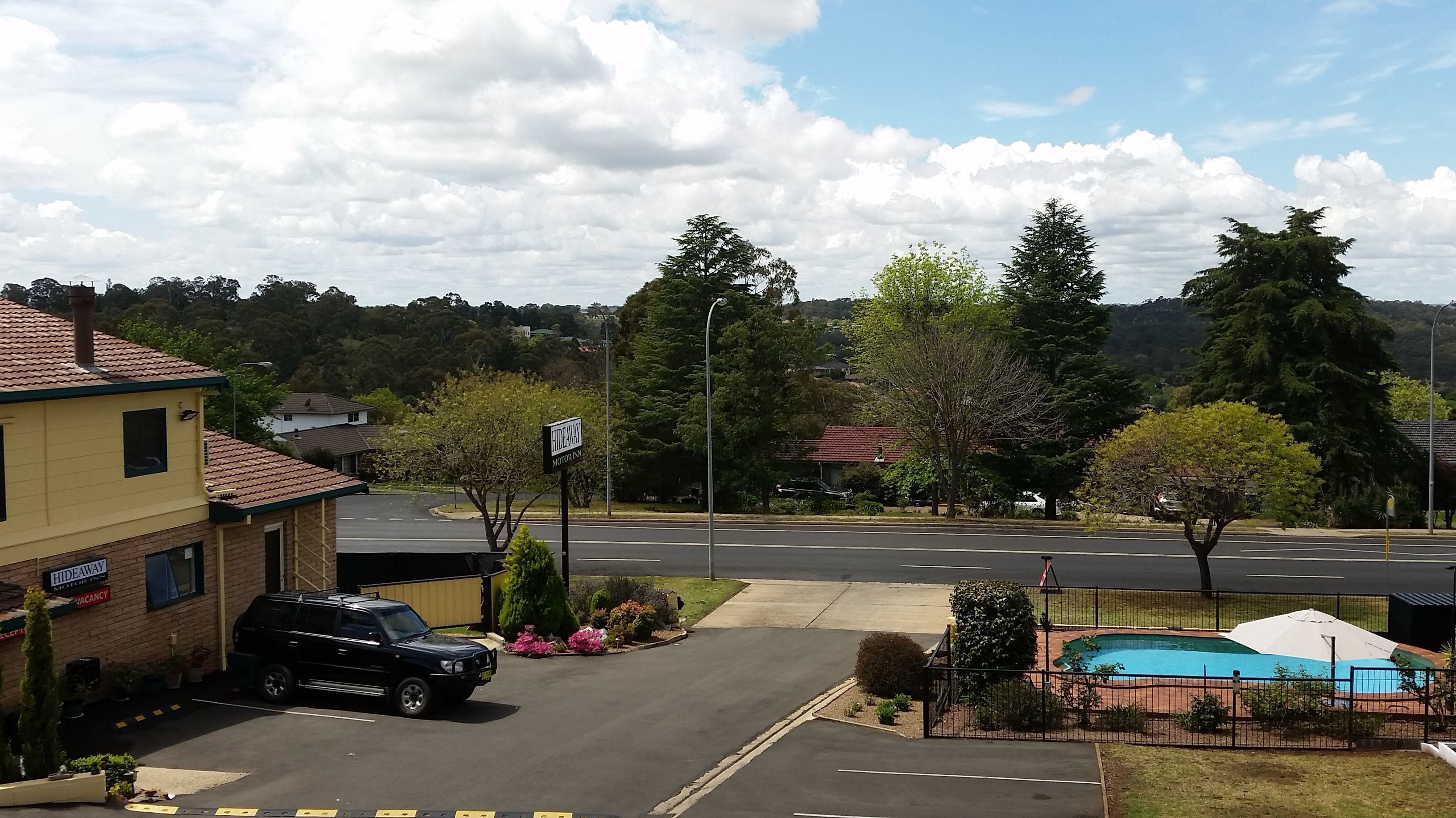 A parking lot with a black SUV, a pool area with a fence, and a building labeled "Hideaway Motor Inn" with a sign indicating a vacancy.