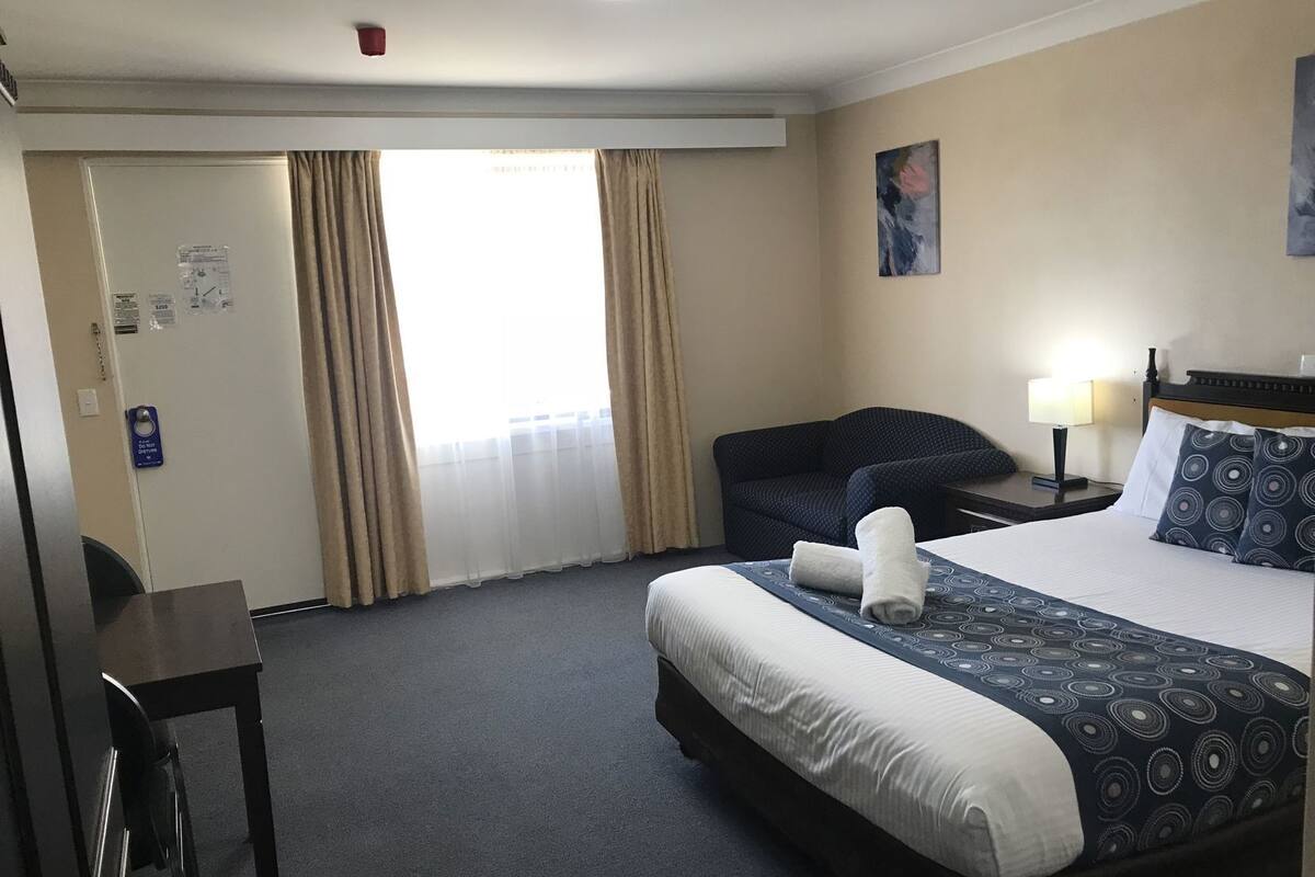 A hotel room with a bed, a desk, a chair, a nightstand, and a window with curtains.