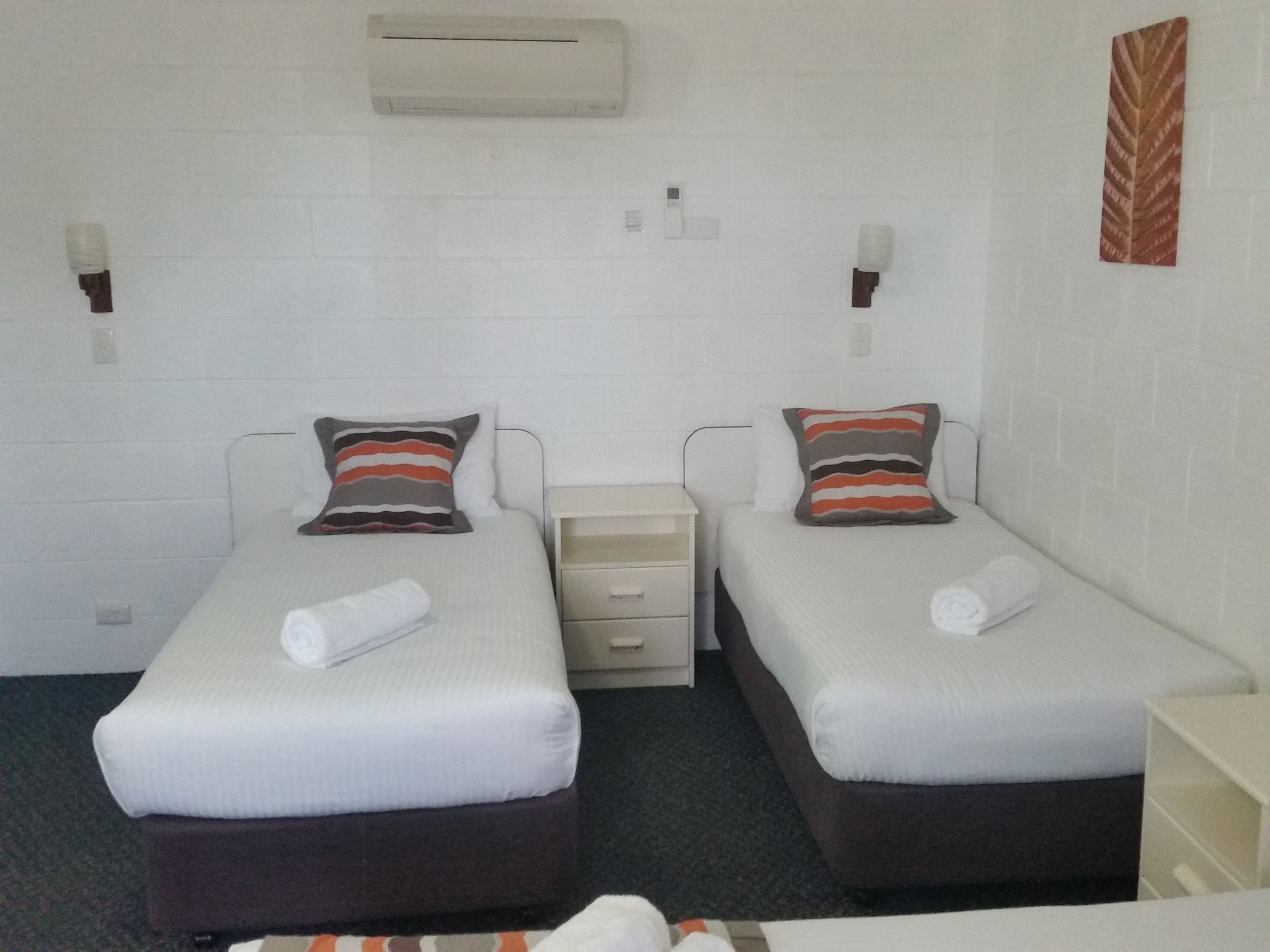 A twin room with two beds, each with a pillow and a rolled towel. There is a small bedside table and a wall-mounted air conditioner.