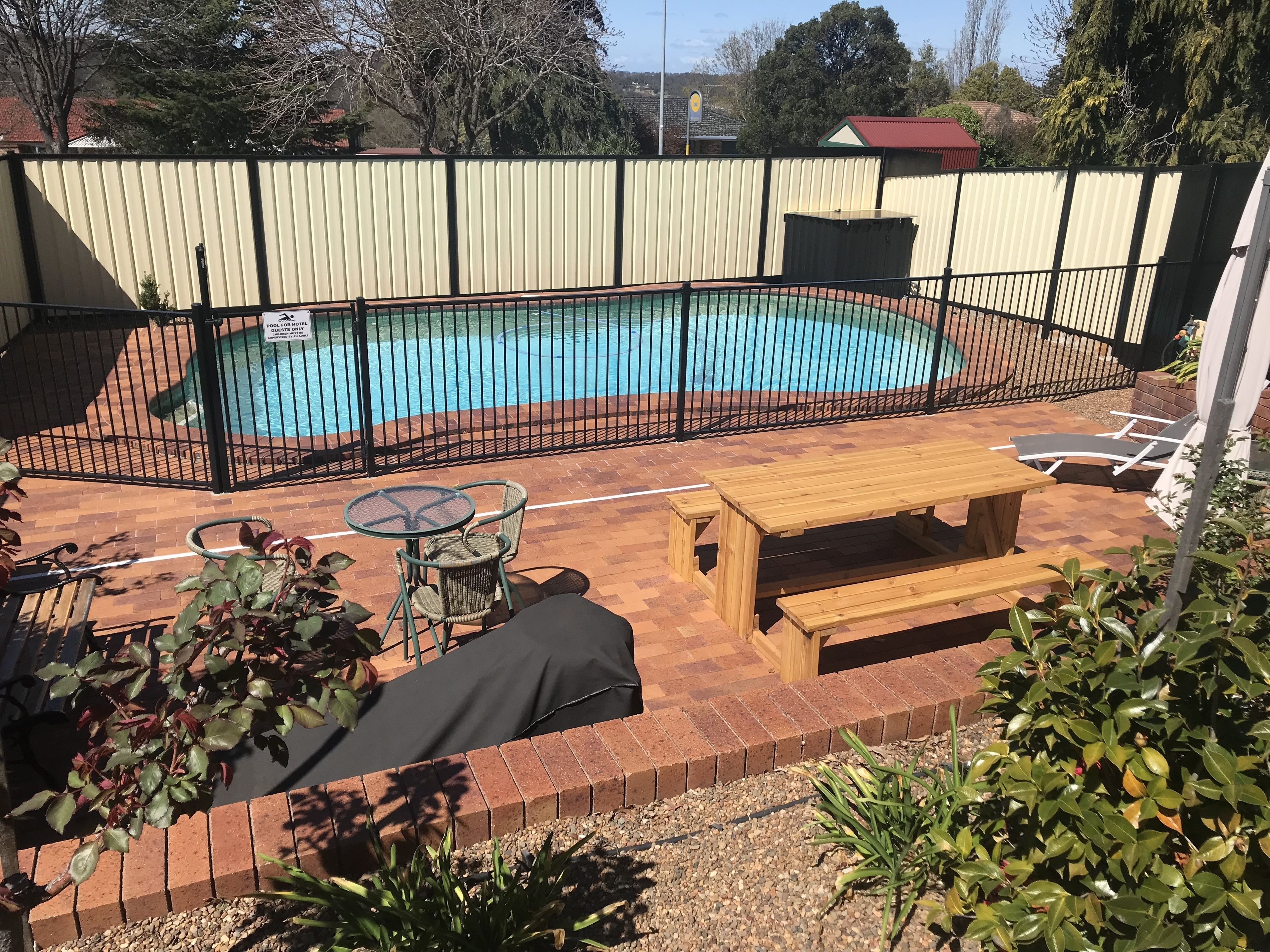 A swimming pool surrounded by a black metal fence, with a wooden bench and a glass table with chairs nearby.