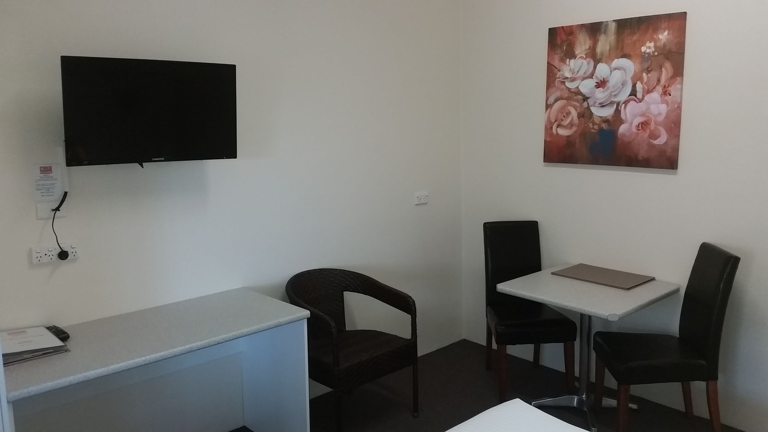 Een kleine kamer met een bureau, twee stoelen, een tafel, een aan de muur bevestigde televisie en een schilderij.