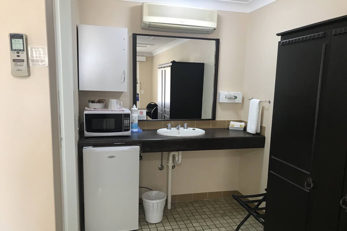 A compact hotel bathroom with a white sink, a microwave, a small refrigerator, a trash bin, and a black cabinet.