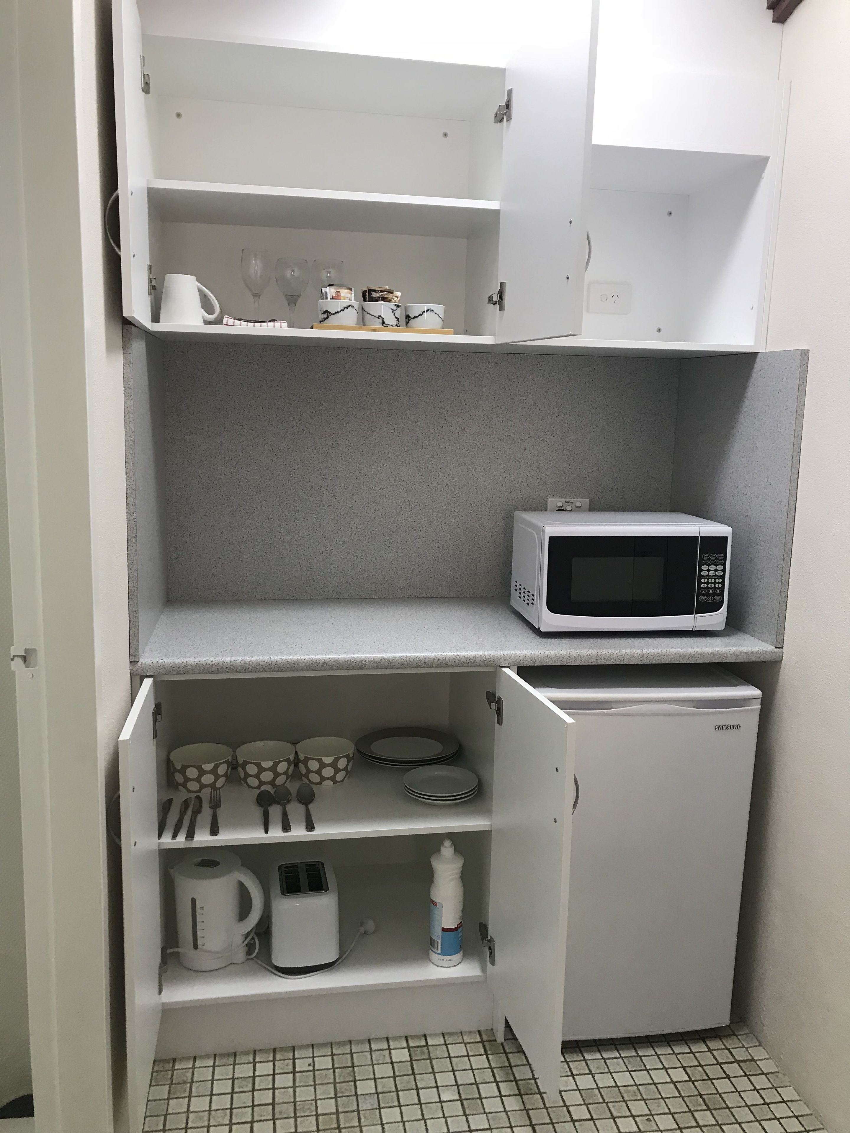 A compact kitchenette with a microwave, a small refrigerator, and shelves holding kitchenware and a kettle.