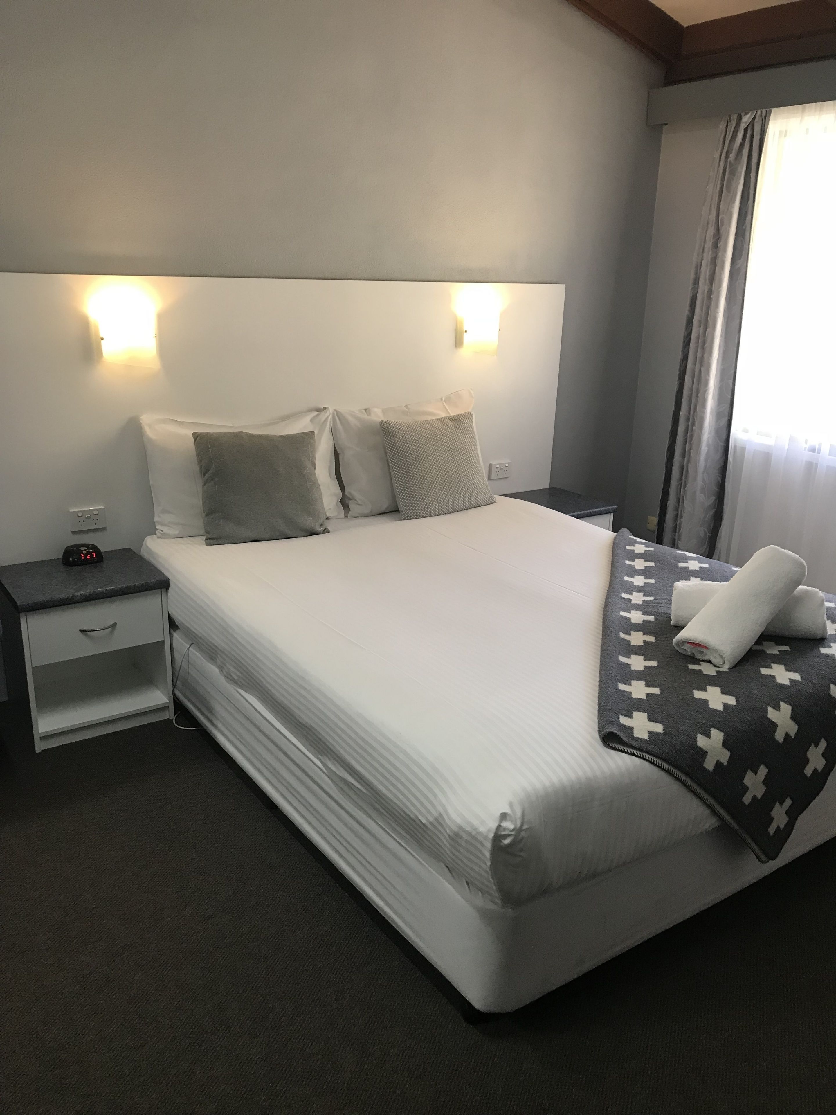 A neatly made bed with a patterned blanket, a bedside table with a clock, and wall-mounted lights.