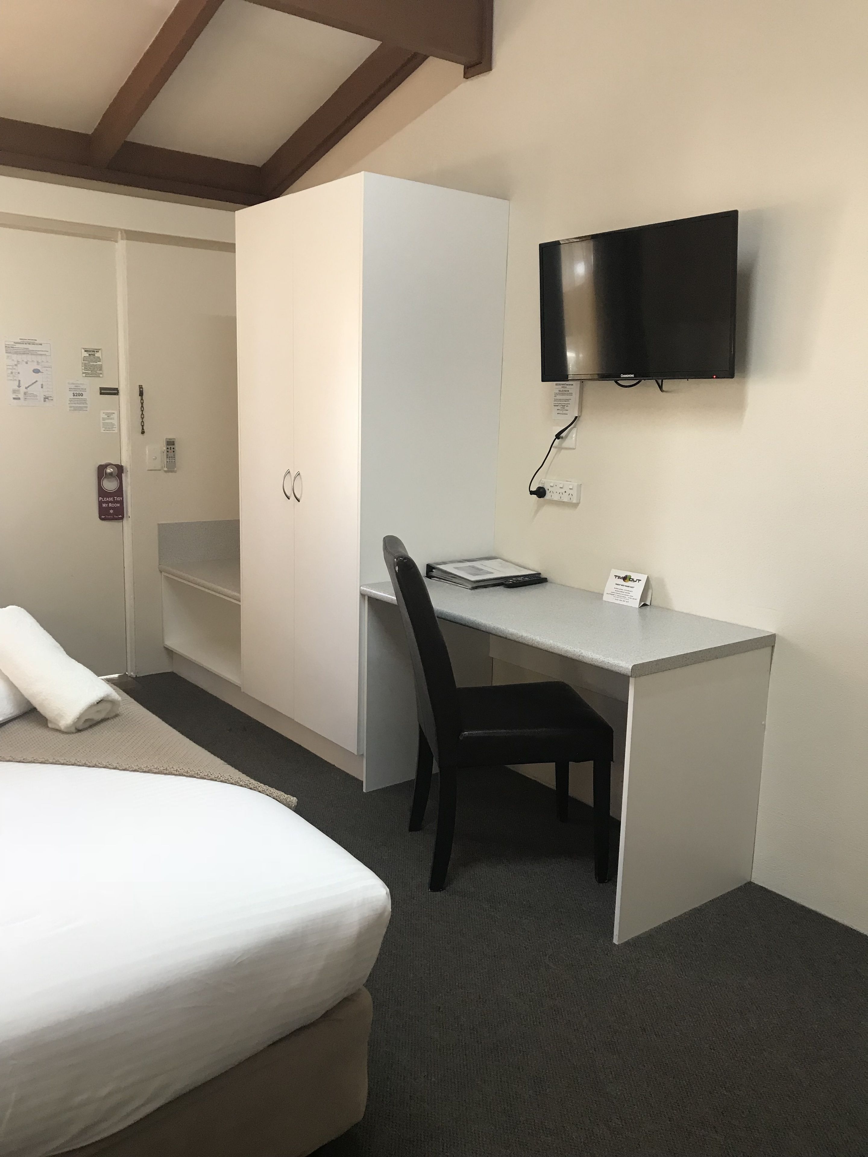 A hotel room with a bed, a desk with a chair, a television mounted on the wall, and a wardrobe.