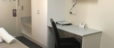 A hotel room with a bed, a desk with a chair, a television mounted on the wall, and a wardrobe.