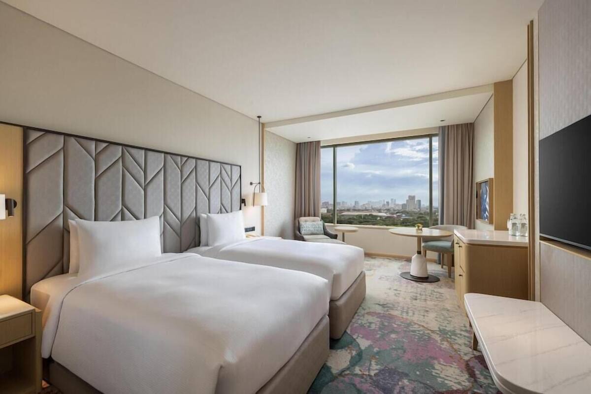 A hotel room with two beds, a desk, a chair, a TV, and a large window with a city view.