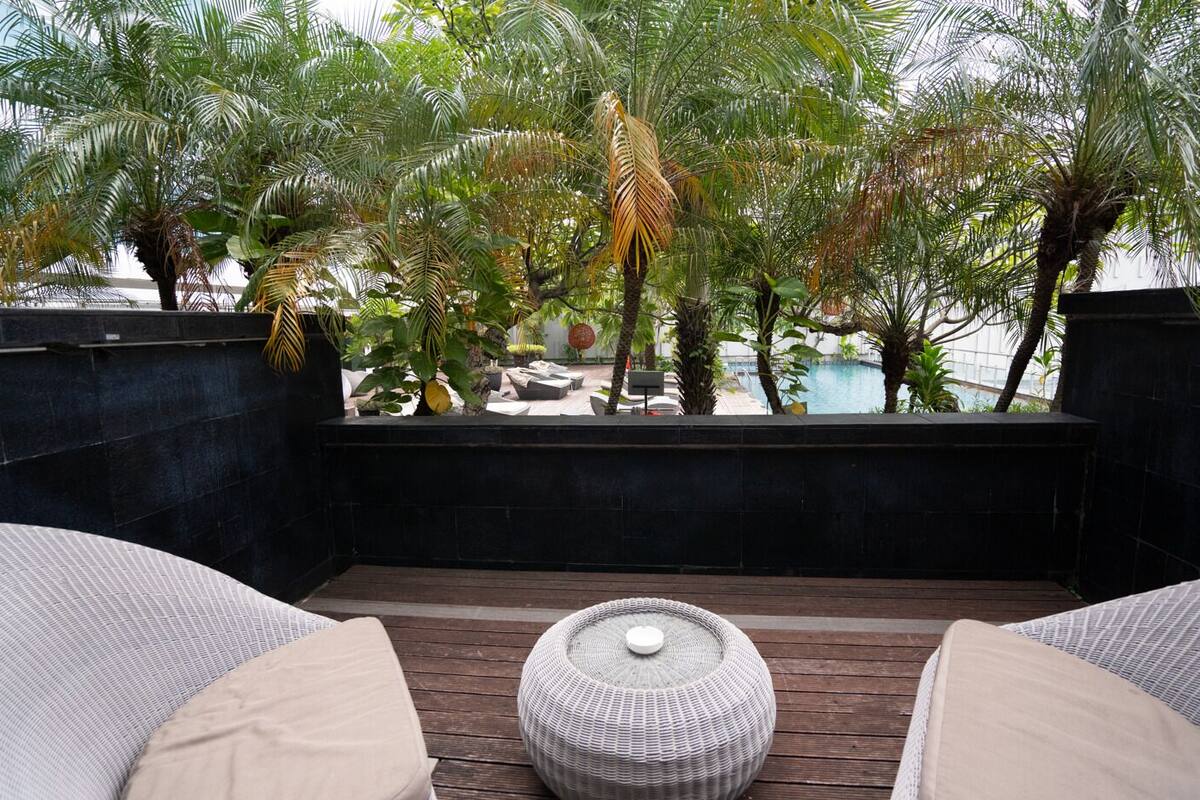 A rooftop terrace with a wooden deck, two cushioned seating areas, and a view of a pool and palm trees.