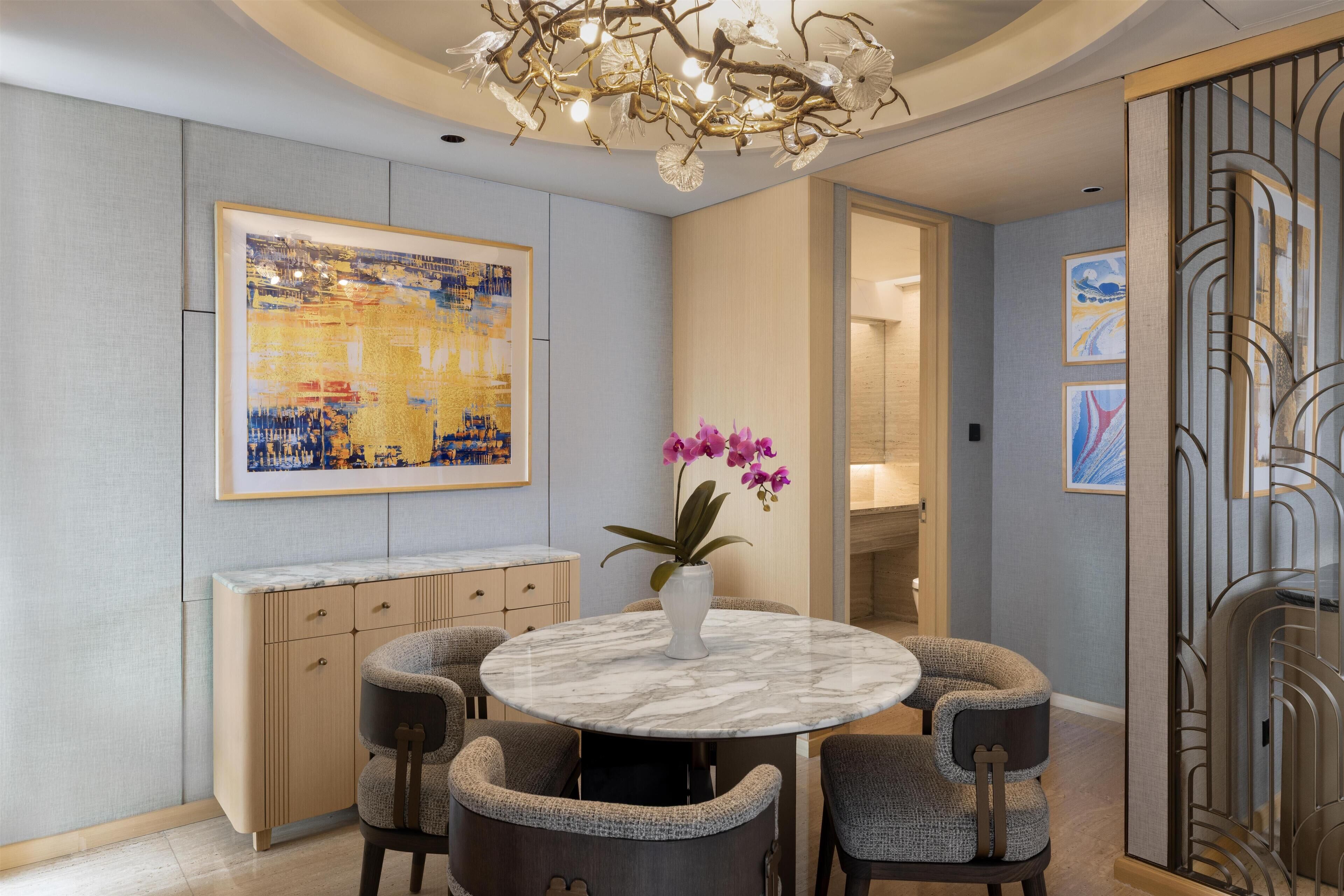 A dining area with a marble table, cushioned chairs, a vase with pink flowers, and a large abstract painting on the wall.