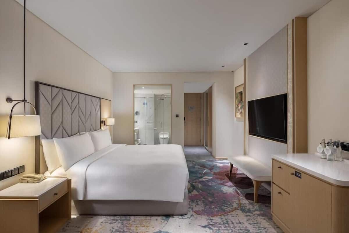 A hotel room with a bed, bedside table, lamp, TV, and a bathroom visible through an open door.