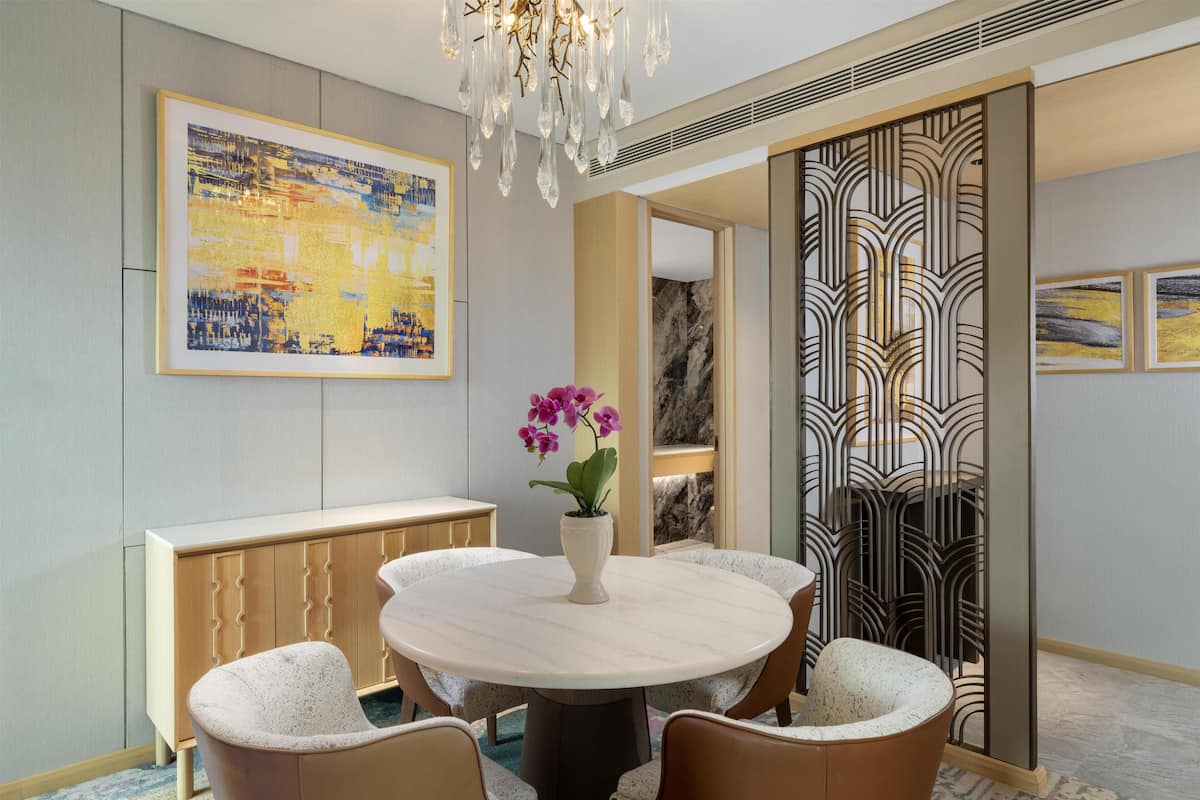 A dining area with a round table, chairs, a vase with flowers, a framed painting, and a decorative screen.