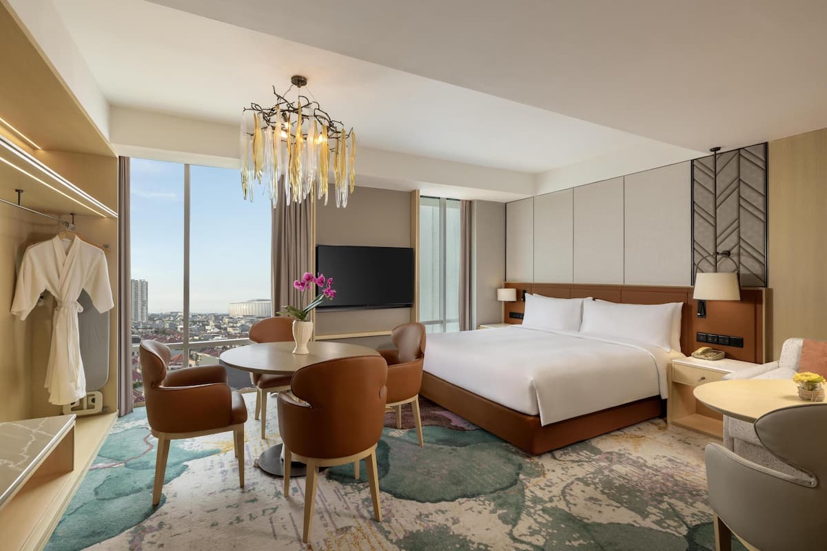 A hotel room with a large bed, a desk with a chair, a small table with a chair, a TV, and a view of the city.