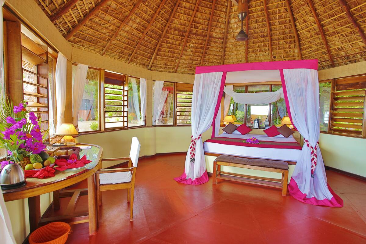A room with a bed, a table with fruits, a chair, and a window with curtains.