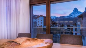 A wooden dining table with a large bowl centerpiece, a window with a view of a mountain, and a balcony with buildings. - Haus Colosseo by Zermatt Premium Apartments (Zermatt)