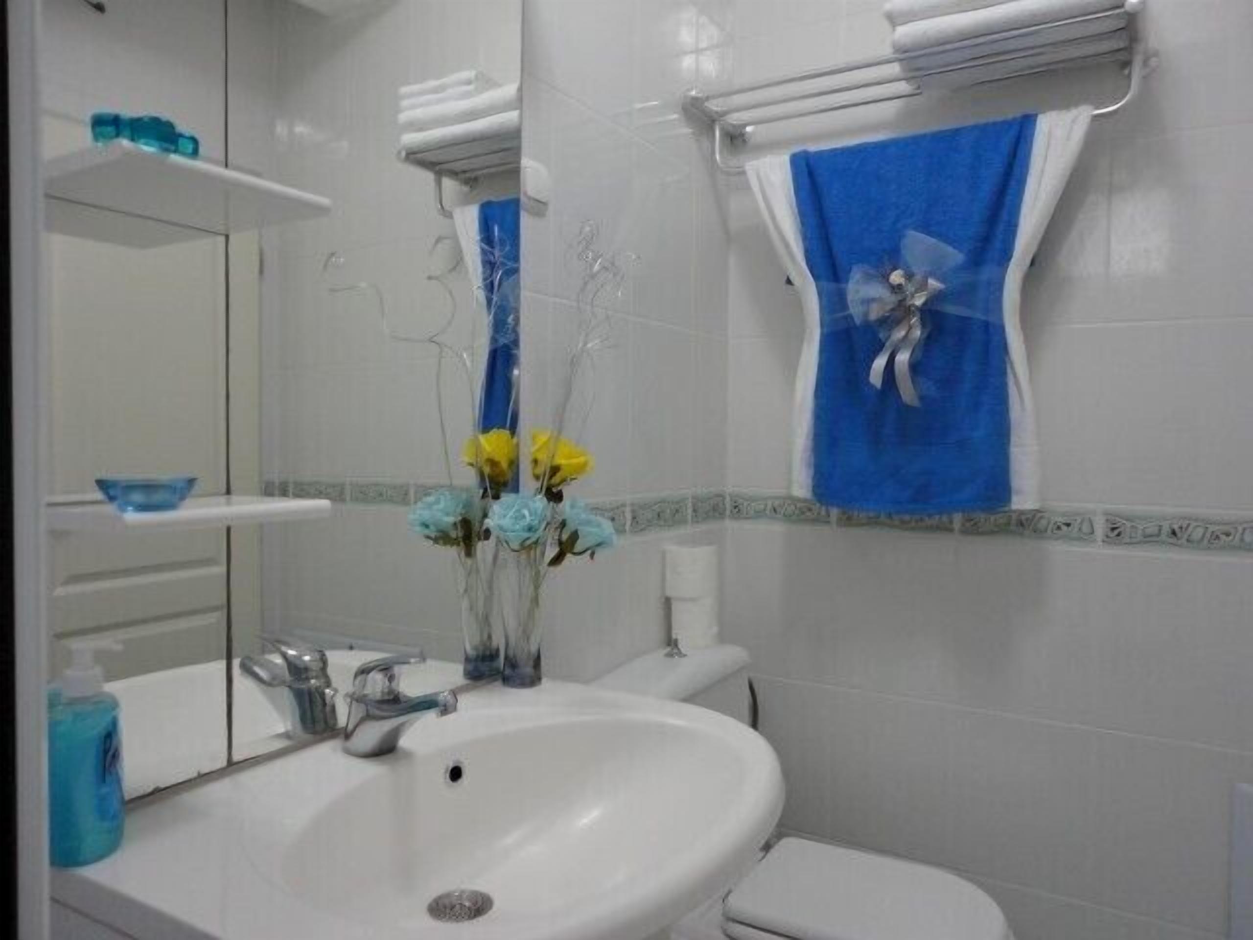 Une salle de bain avec un lavabo blanc, un miroir, une serviette bleue et un vase rempli de fleurs.