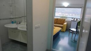 A compact hotel room with a bathroom featuring two sinks and a shower. The room includes a small sofa and a dining table with a view through a window. - Accommodation J&T (Ohrid)