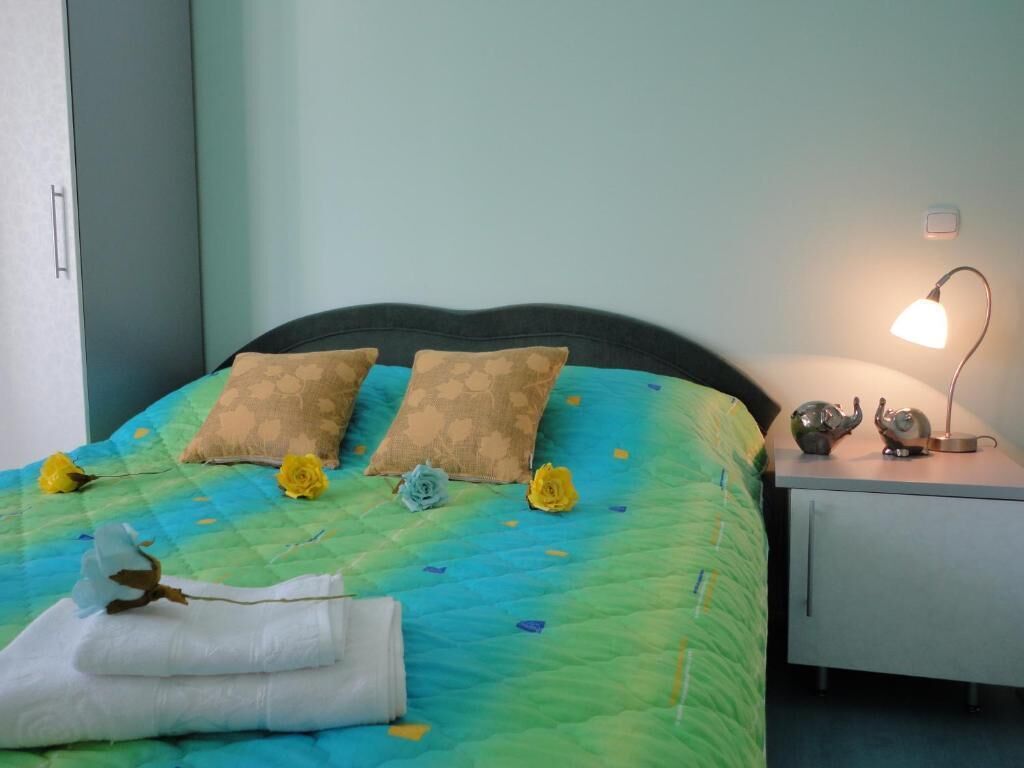 Ein ordentlich bezogenes Bett mit einer gesteppten Decke, zwei Kissen und Zierblumen. Eine Nachttischlampe und ein kleiner Tisch mit Teekanne sind ebenfalls zu sehen.