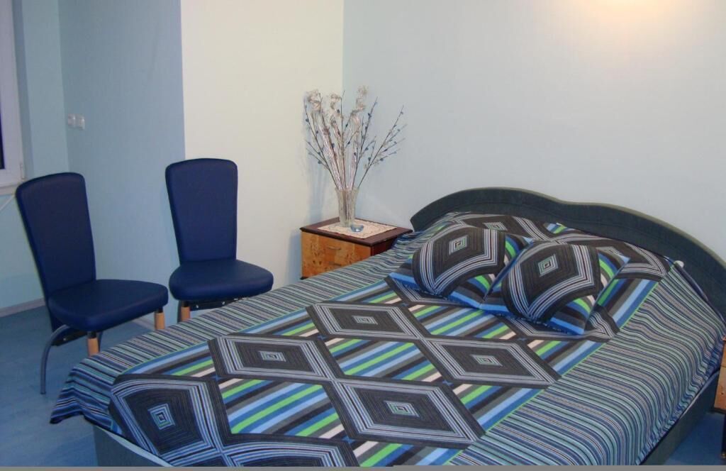 A bedroom with a bed, two blue chairs, a nightstand, and a vase with dried branches.