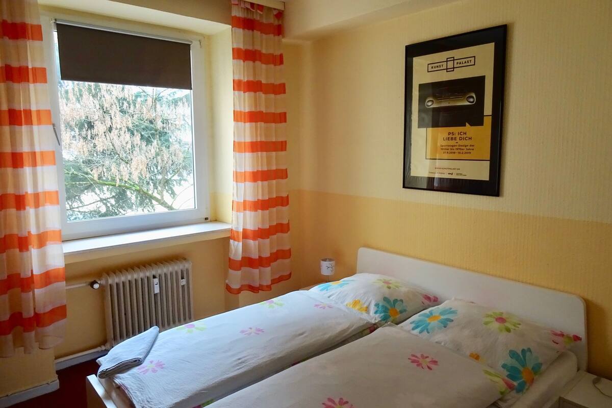 A bedroom with two beds, a window with curtains, a radiator, and a framed picture on the wall.