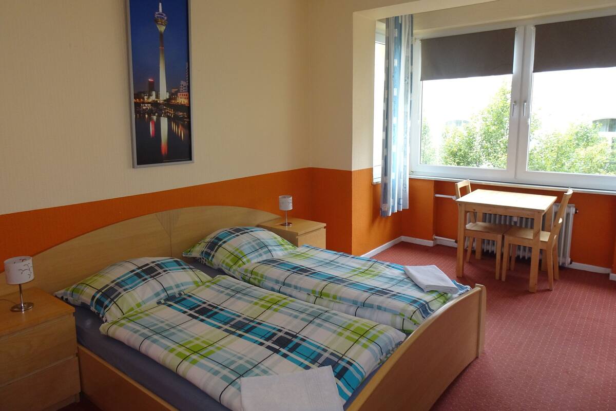 A hotel room with a bed, a desk, and a chair. There is a window with curtains, a radiator, and a poster on the wall.