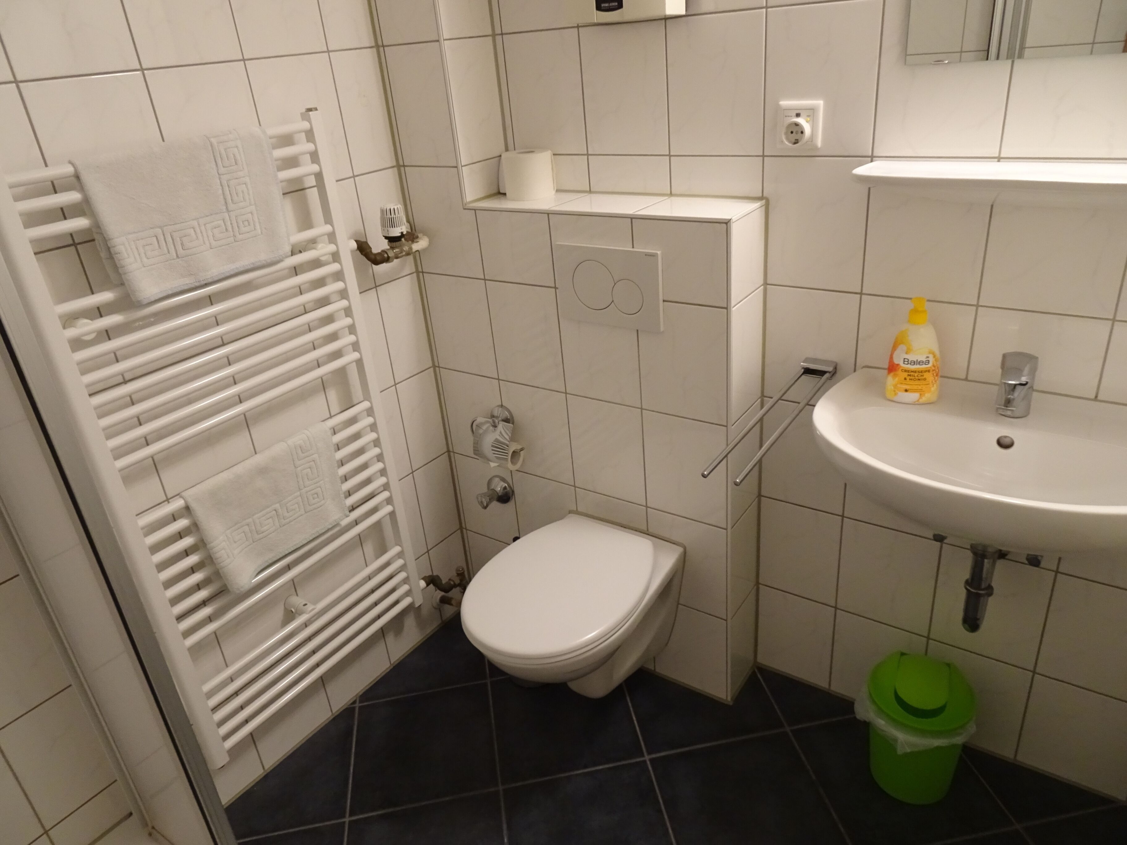 A small bathroom with a toilet, a sink, a towel rack, and a green trash bin.