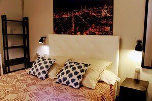 A hotel room with a bed, two pillows, a nightstand with a lamp, and a cityscape view on the wall. - The Buba House - Hostel (Barcelona)
