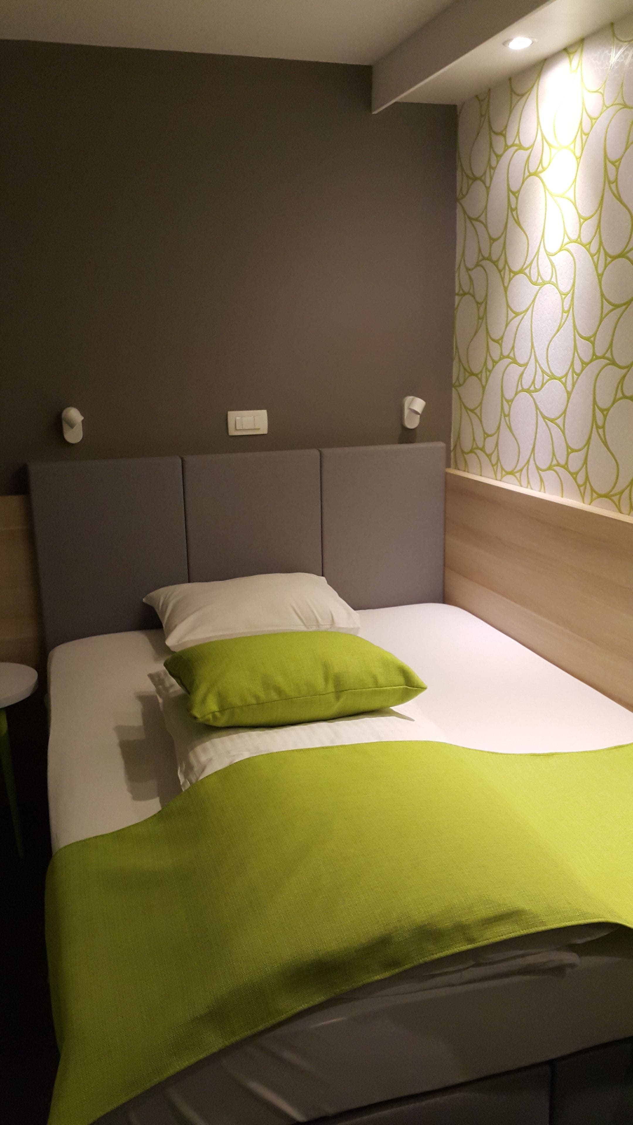A neatly made bed with a green pillow and blanket, a grey headboard, and a wall with a green and white pattern.