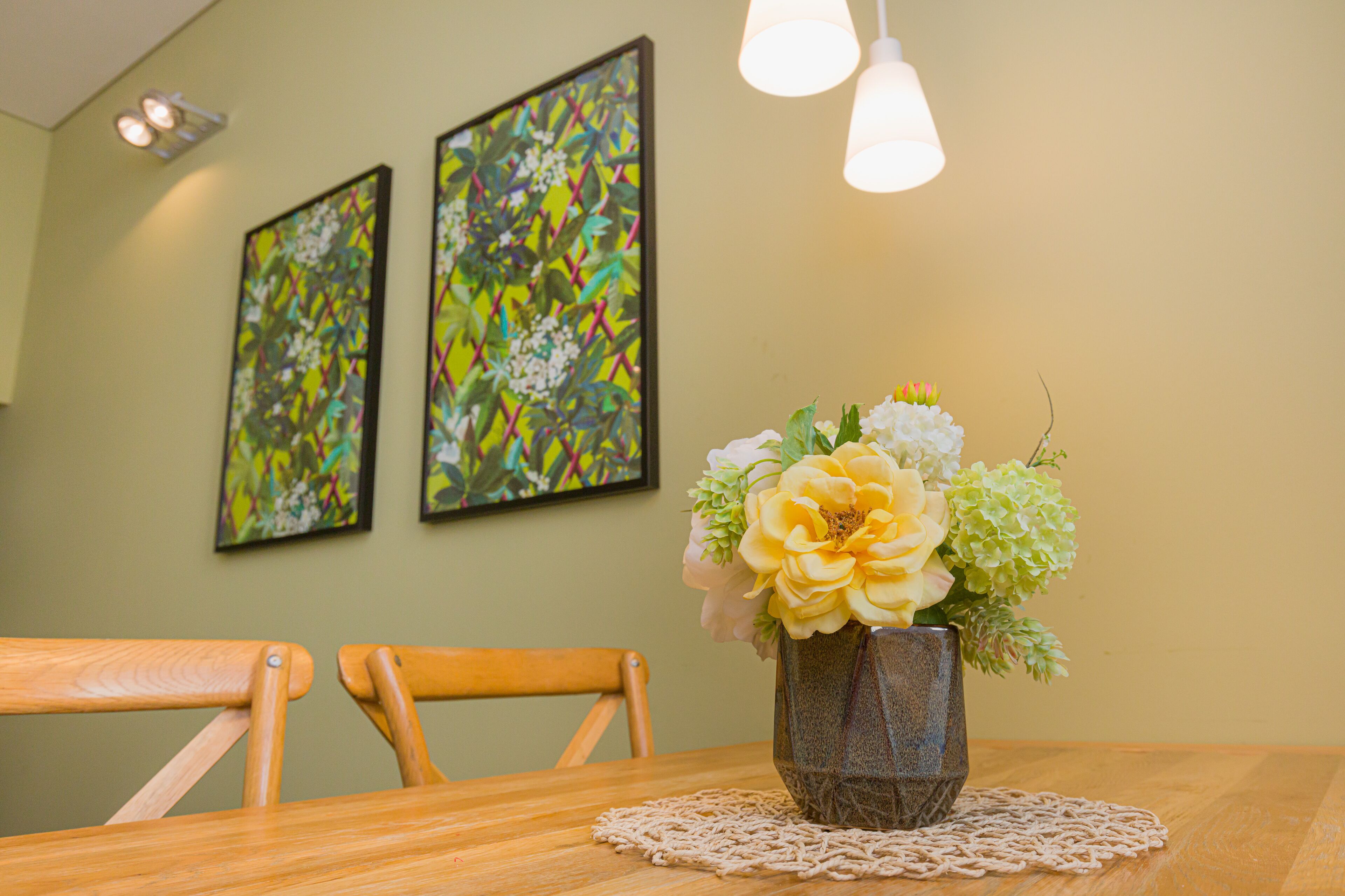 A dining table with a vase of flowers, two framed artworks on the wall, and a wooden chair.