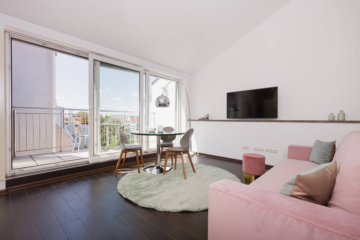 A modern living room with a glass door leading to a balcony, a flat-screen TV mounted on the wall, and a pink sofa.