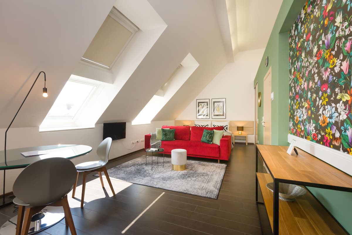 A modern loft apartment with a high ceiling, skylights, a red sofa, a glass dining table, and a colorful floral wall.