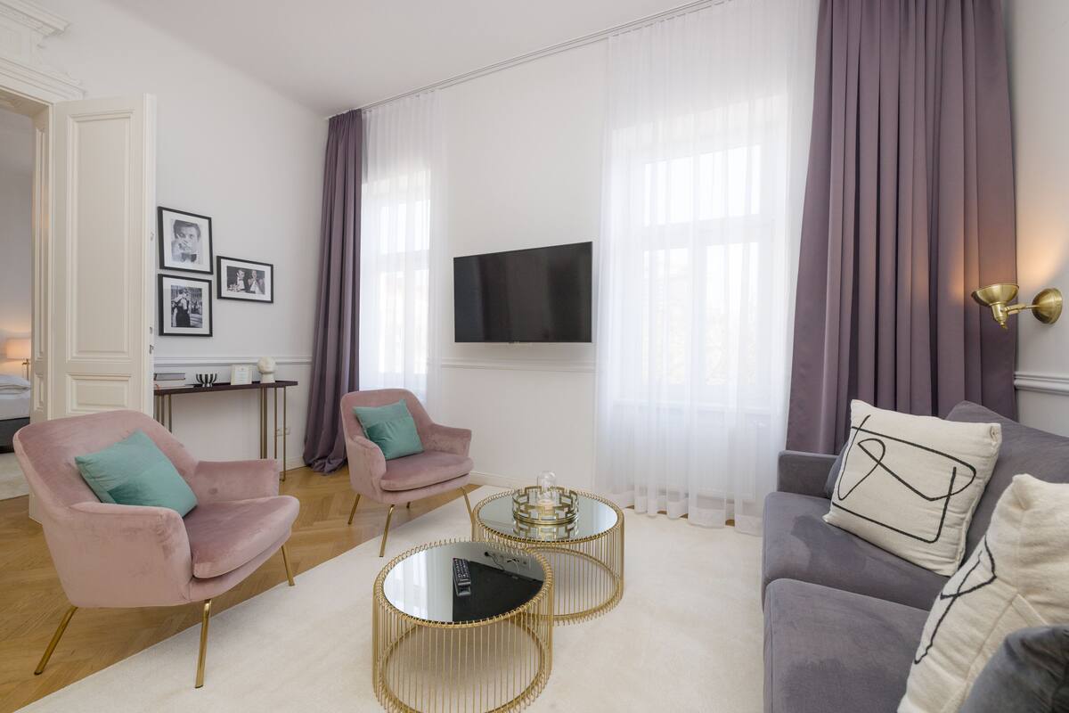 A modern living room with a grey sofa, two pink armchairs, a coffee table, and a TV mounted on the wall.