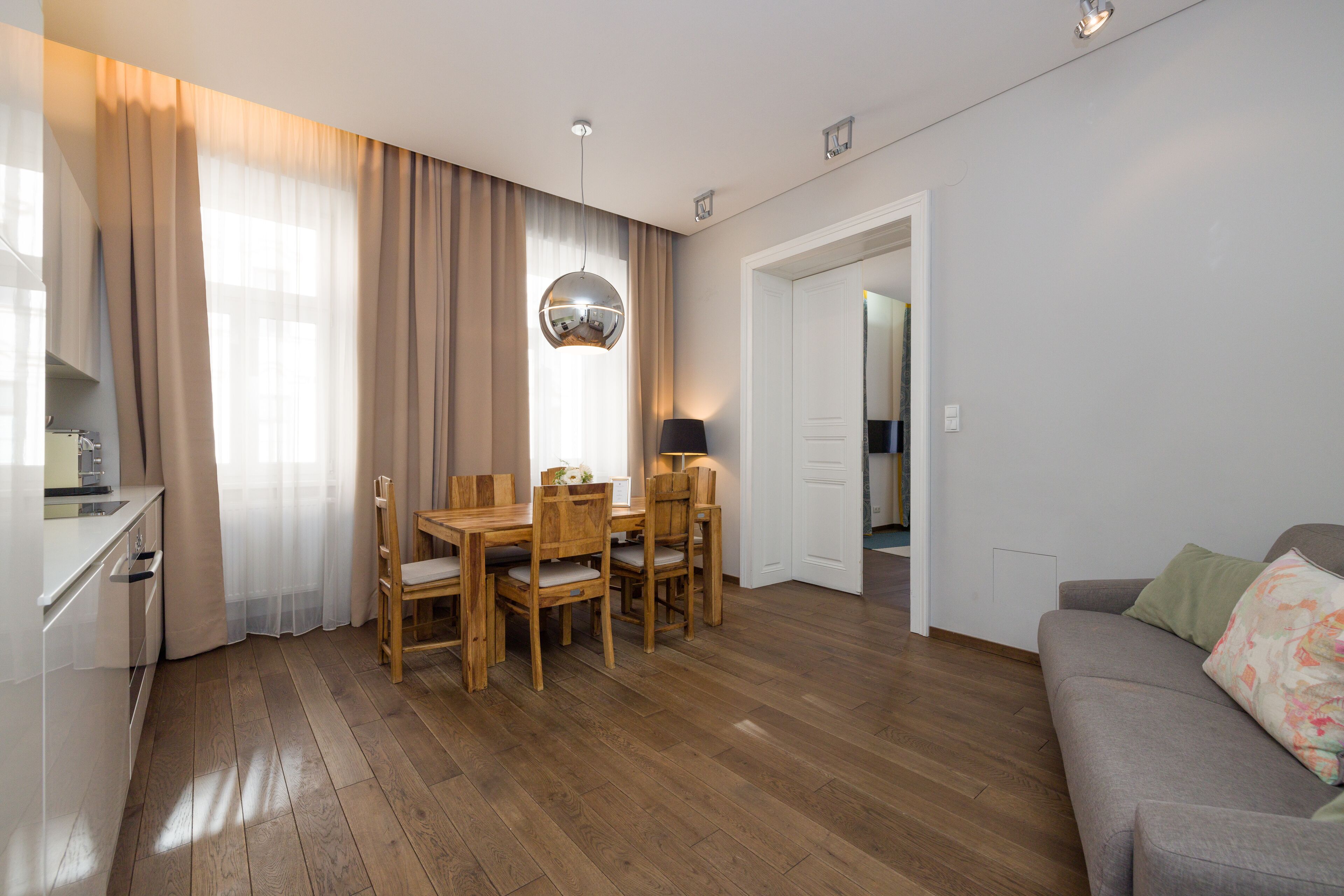 Een moderne woonkamer met een houten eettafel, stoelen, een grijze bank en een keuken met een wit aanrecht en kastjes.