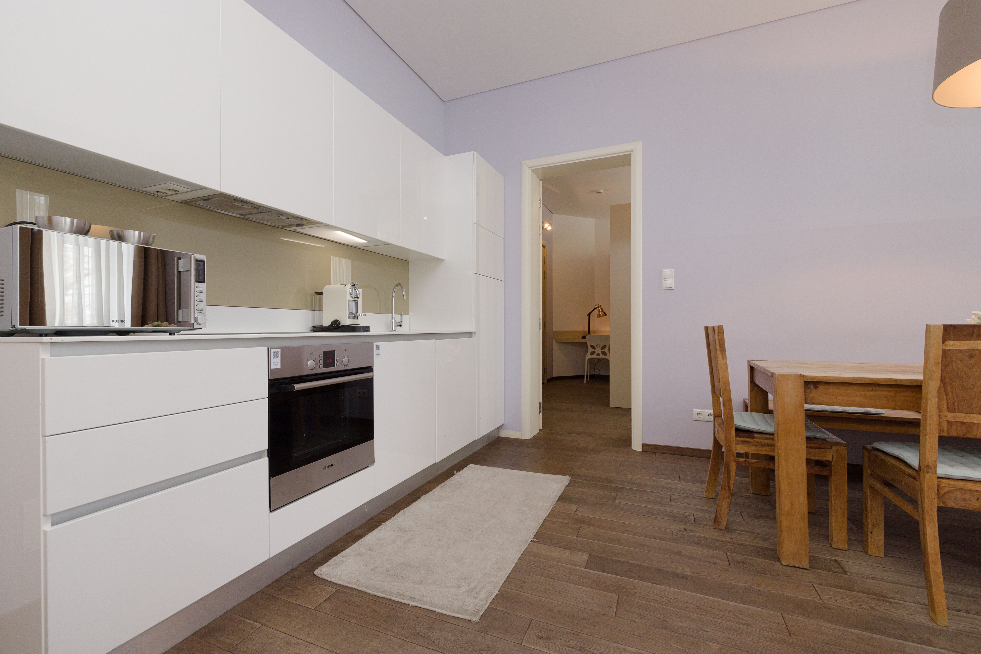 A modern kitchen with white cabinets, a built-in oven, and a coffee machine. There is a dining area with a wooden table and chairs.