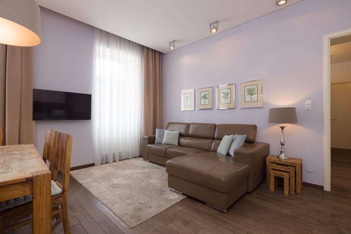 A living room with a brown leather sofa, a wooden dining table, a TV mounted on the wall, and framed pictures on the wall.