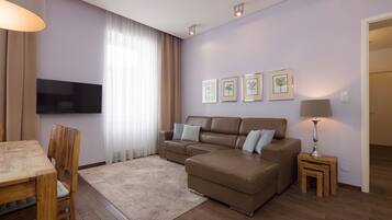 A living room with a brown leather sofa, a wooden dining table, a TV mounted on the wall, and framed pictures on the wall.