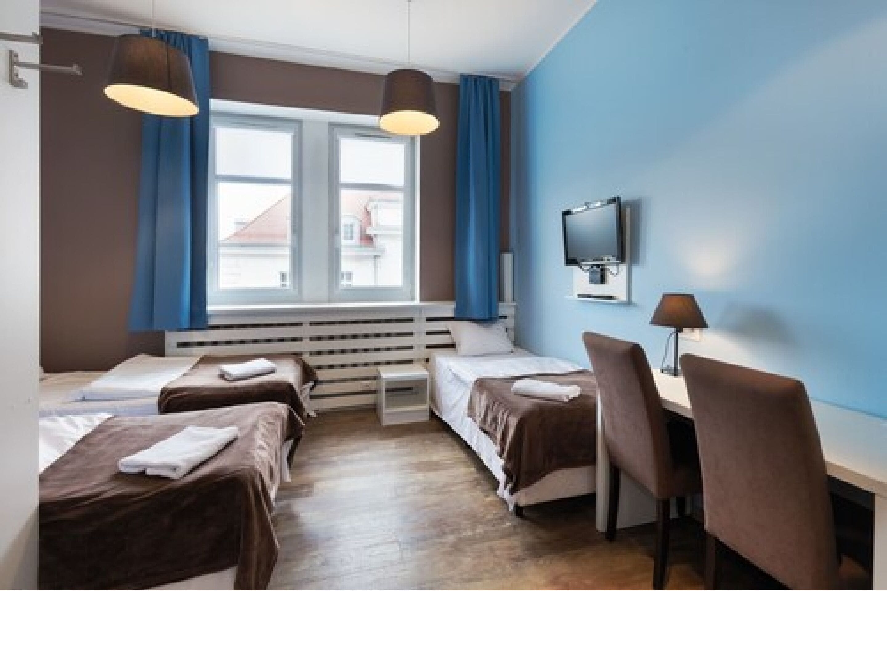 A hotel room with two single beds, a desk, and a chair. There is a window with curtains, a flat-screen TV mounted on the wall, and a heater under the window.