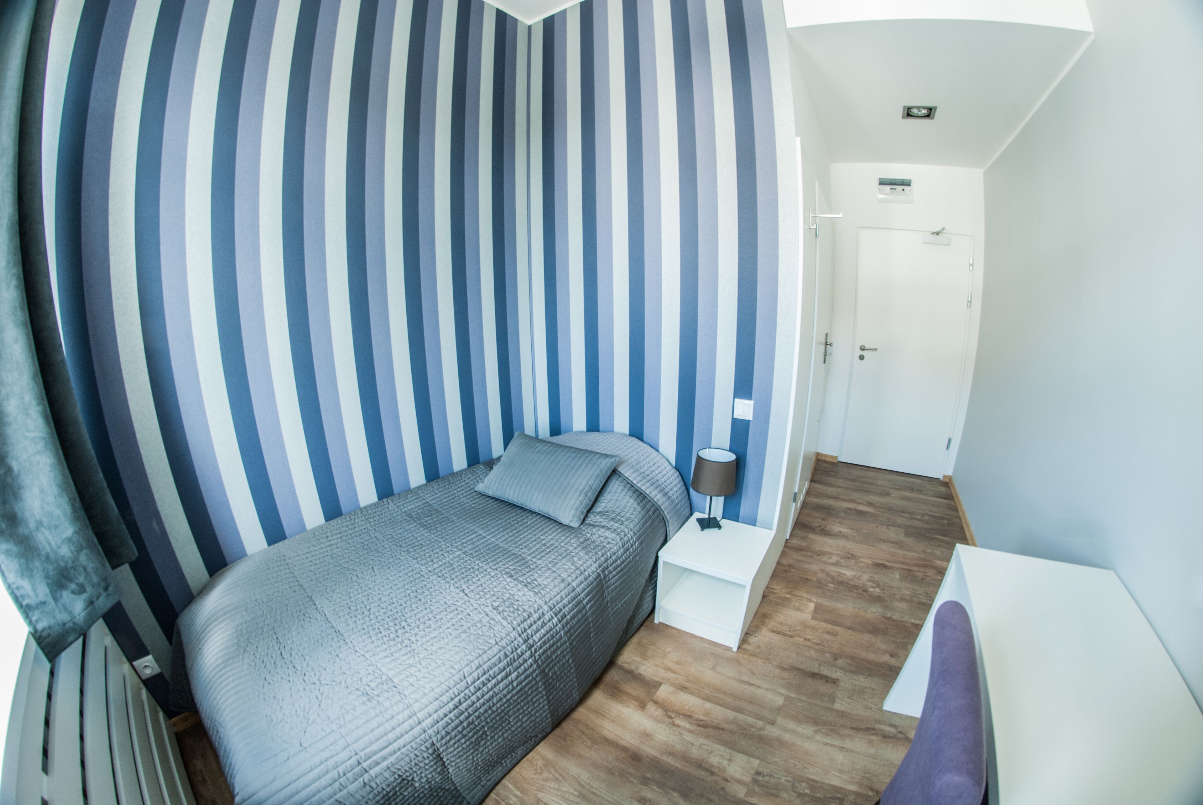 Un dormitorio con pared a rayas, una cama, una mesita de noche con lámpara y una puerta.