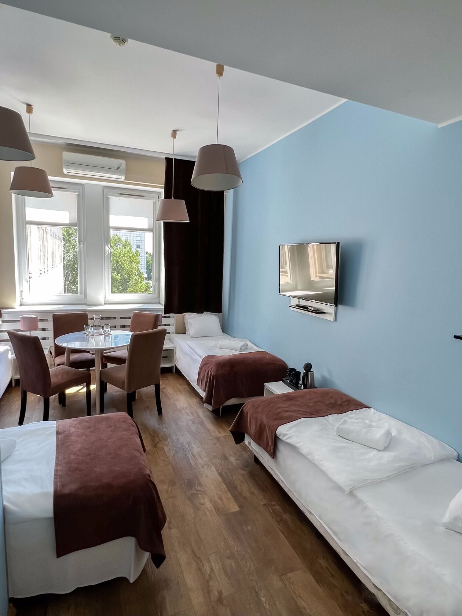 Ett hotellrum med två sängar, ett matbord med stolar, en tv på väggen och stora fönster med gardiner.