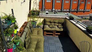 Una terraza en la azotea con zona de estar, plantas en macetas y vista a un edificio con grandes ventanales.
