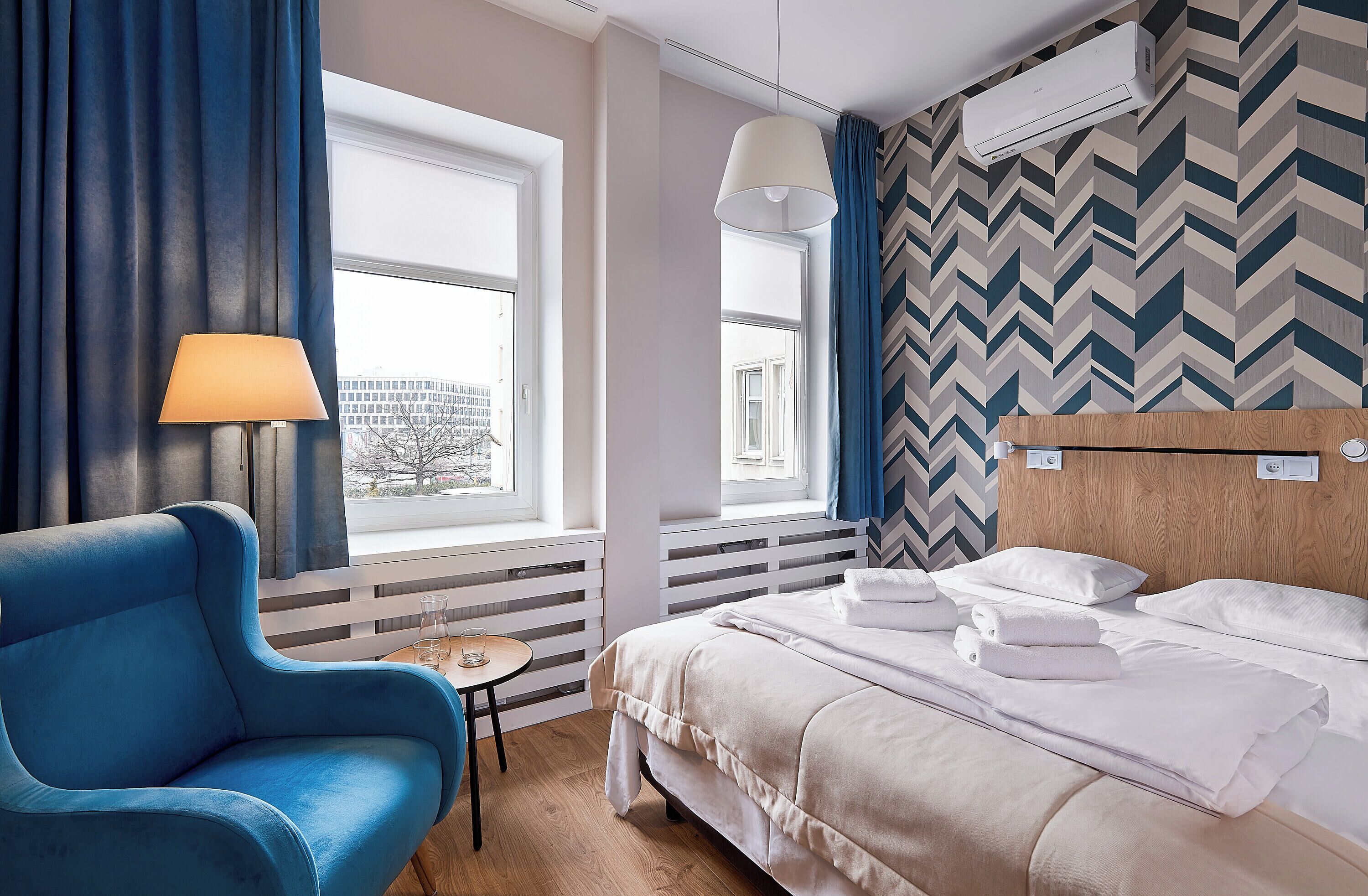 A hotel room with a bed, a blue armchair, a small table, and a window with curtains.