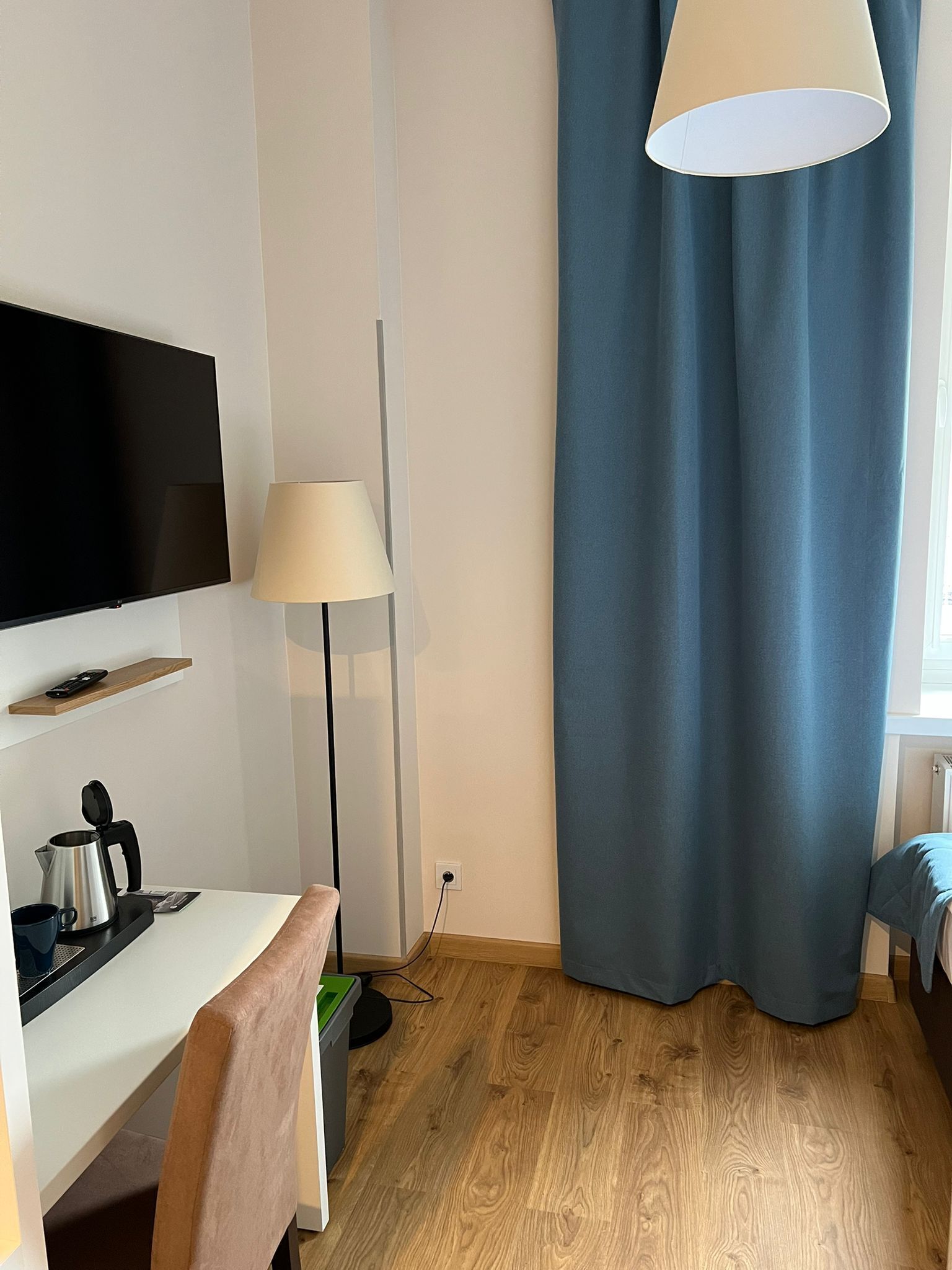 Habitación de hotel moderna con una televisión de pantalla plana, un escritorio con tetera, una lámpara y una cortina azul.