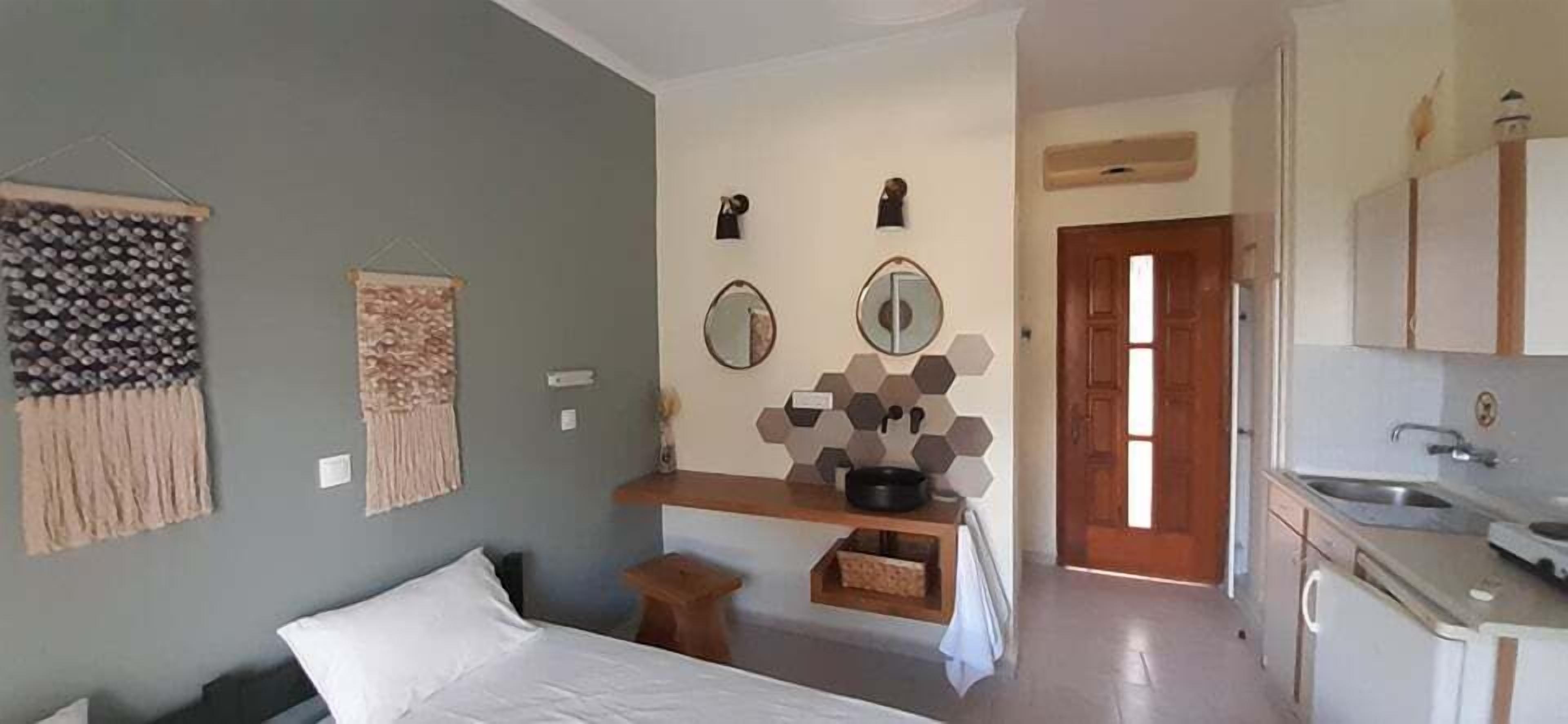 A compact hotel room with a bed, a small stool, a shelf with kitchen items, and a door leading to the kitchen.