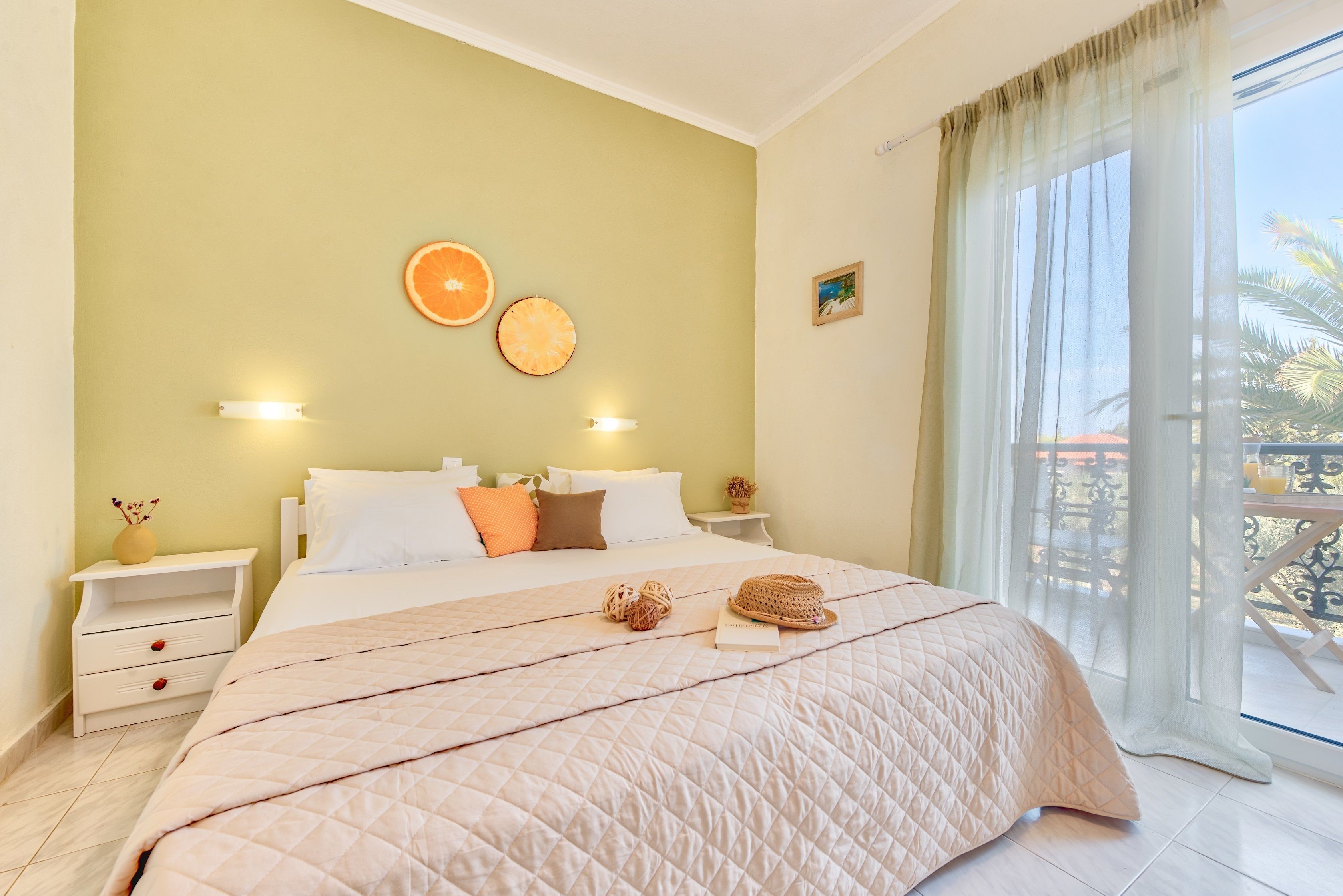A bedroom with a large bed, a nightstand, a wall-mounted lamp, and a window with a view of palm trees.