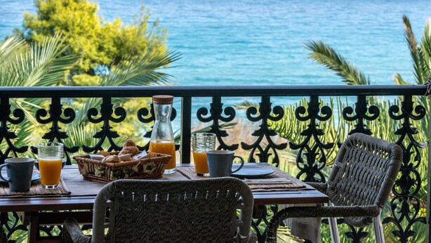 Mesa posta com itens de café da manhã, incluindo um copo de suco de laranja, uma garrafa e uma cesta de doces, com vista para um oceano panorâmico.