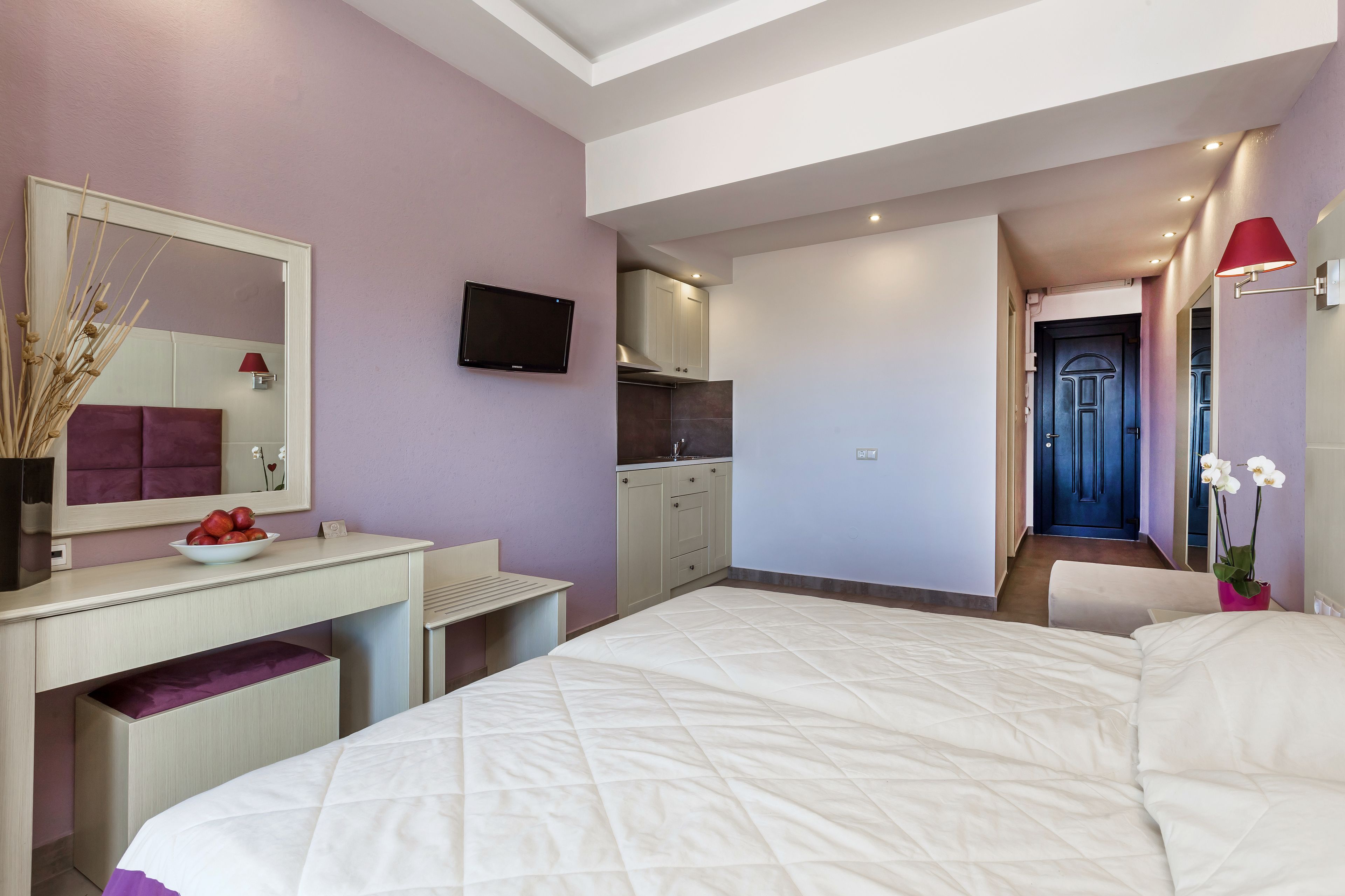 A bedroom with a large bed, a desk with a mirror, a television, and a kitchenette in the background.