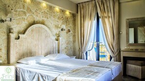 Quarto com parede de pedra, cabeceira de madeira, cama com roupa de cama branca, mesa de cabeceira, cômoda e janela com cortinas.