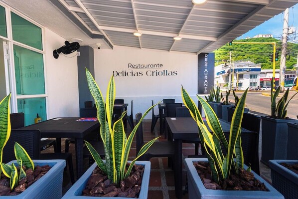 Een terras met tafels en stoelen, plantenbakken en een muur met de naam van het restaurant erop.